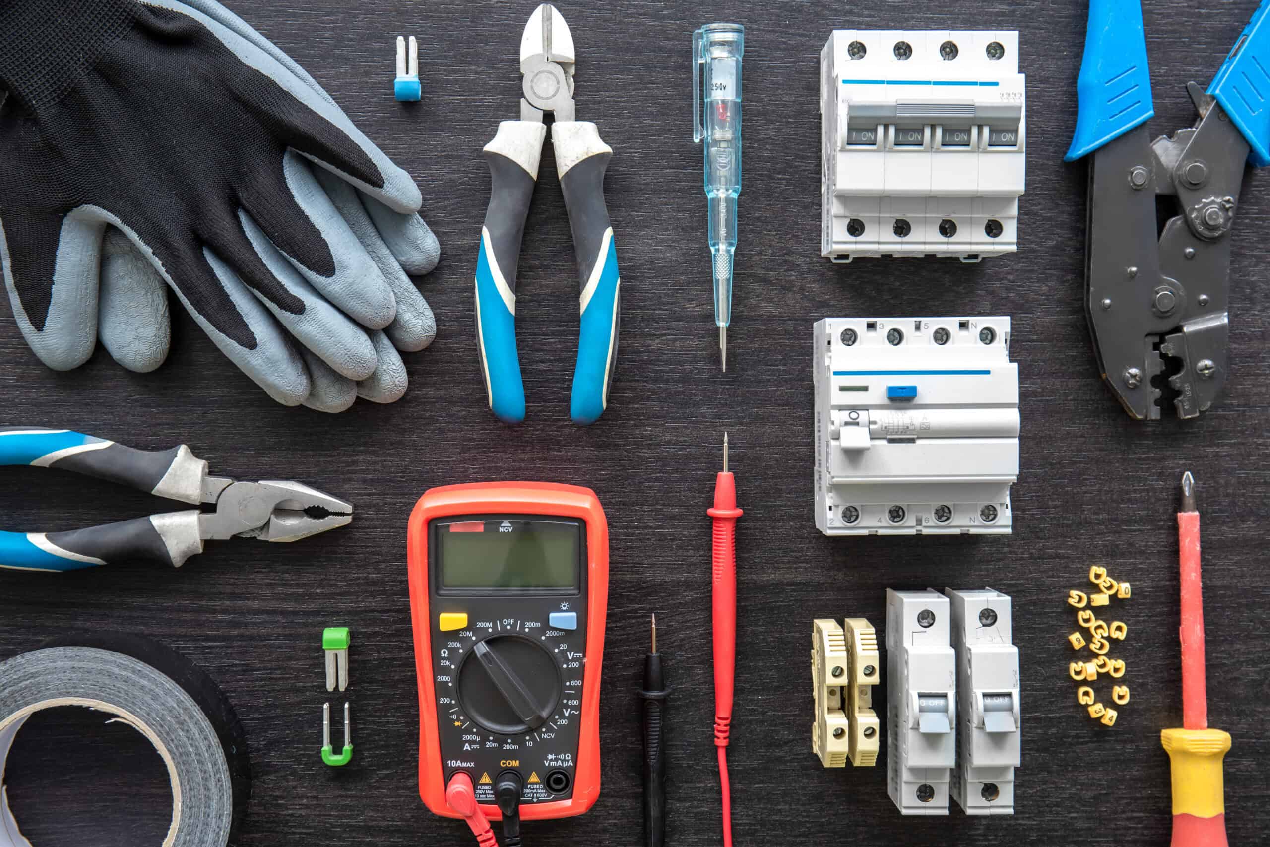 A selection of Electrical Supply For Contractors is neatly arranged on a dark surface, featuring gloves, pliers, a multimeter, screwdrivers, electrical tape, and circuit breakers.