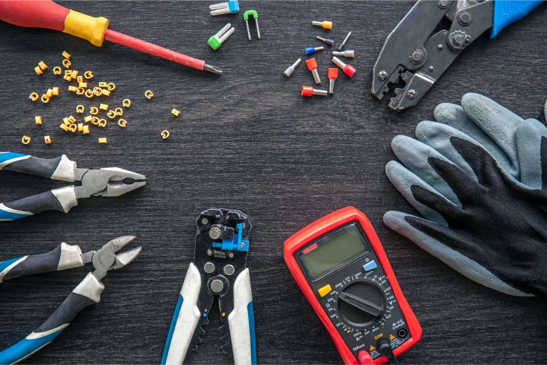 A variety of electrical tools and components, perfect for any home builder's supplies, including pliers, screwdriver, crimping tool, multimeter, gloves, and connectors, are neatly arranged on a dark wooden surface.