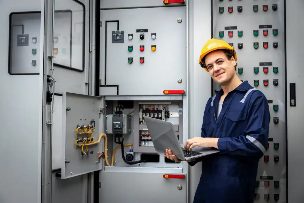 Why Traditional Electrical Systems Fail Modern Buildings 90% of the Time (And How to Fix It) 1 A worker analyses an electrical system in a hard hat and coveralls using a laptop while standing in front of an open electrical control panel with various switches and indicators.
