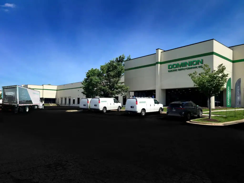 A commercial building with "Dominion Electric Supply Co." signage, several white service vans, a Dominion delivery truck, and a car parked in front under a clear blue sky, showcasing the operations of an Electrical Distribution Company.