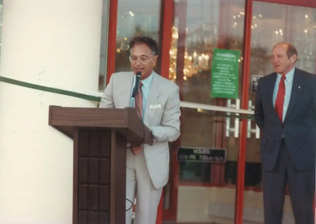 Nuestra historia 4 A man in a light suit speaks at a podium outside an Electrical Distribution Company building, while another man in a dark suit stands nearby. Glass doors and signage are visible in the background.