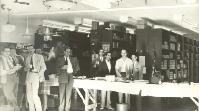 Nuestra historia 1 A group of people in suits and dresses stand around a long table with food and drinks in a store or office with shelves in the background.